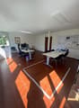 Large open office space with hardwood floors, white walls, and modern furniture including desks, chairs, and a whiteboard. The room has good natural lighting from the windows.