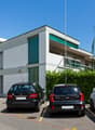 A modern, multi-story building with a gray exterior and green accents. There is a parking lot in front of the building with several cars parked.