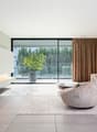Spacious living room with large windows offering a view of the surrounding greenery. The room features a modern and minimalist design, with a sleek TV console, a comfortable sofa, and a stylish armchair. The curtains add a touch of warmth to the space.