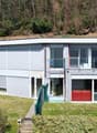 Two-story modern house with a flat roof, large windows, and a balcony or terrace. The exterior is a combination of white and red panels, with some stone accents. The house is surrounded by a grassy yard and trees.