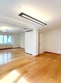 This image shows a spacious, open-plan room with hardwood floors and large windows. The room has a modern, minimalist design with a long, rectangular light fixture hanging from the ceiling. The walls are painted a neutral color, and there is a door visibl