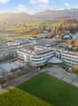The image shows a large multi-story building complex surrounded by a grassy area and trees. The buildings have a modern, industrial design with flat roofs and large windows. The complex appears to be situated in a hilly, mountainous area with a town visib