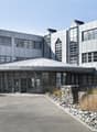 Large modern commercial building with a glass and metal facade, a domed roof structure, and a paved parking area in the foreground. The building has multiple floors and large windows. There is also a gabion wall feature with stones near the entrance.