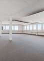 Large open-plan office space with high ceilings, white walls, and large windows providing natural light. The room has a carpeted floor and several support columns. The space appears to be unoccupied and ready for use.