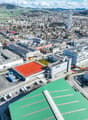 Large industrial area with multiple buildings, some with red tennis courts, multiple solar panels, extensive parking spaces, and multiple cranes and construction vehicles