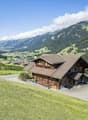 Mountain chalet, wooden exterior, pitched roof, front porch, driveway, green fields, mountain range, clear skies