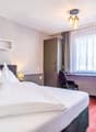 Spacious bedroom with a large bed, white bedding, and a desk area. The room has large windows with curtains, providing natural light. There are shelves on the wall and a decorative plant. The room has a modern and cozy feel.