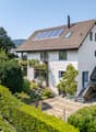 2 story house, brown roof, solar panels, white walls, surrounded by trees and shrubs, balcony
