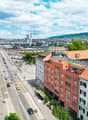 The image shows a panoramic view of a city skyline. There are tall modern buildings, including a green glass skyscraper, as well as older red-roofed buildings. The city is surrounded by hills and mountains in the background. The streets are busy with traf