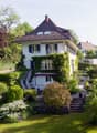2 story house, white exterior, brown roof, balcony, surrounded by greenery