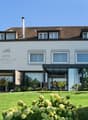 4-star Hotel Burgblick, 3-story building with a brown tiled roof, large windows, and a glass-enclosed patio area. The hotel is surrounded by a well-manicured lawn and lush landscaping, including flowering plants and shrubs.