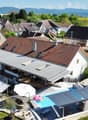 Two-story house with brown roof, solar panels, outdoor swimming pool, patio with patio umbrellas, garden, nearby trampoline, and distant views of mountains.