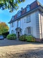 3 story house, white exterior, raised ground floor, gravel driveway, entrance lamps, car parked, garden with shrubs, trees