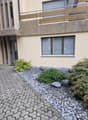 paved driveway, balcony with metal railing, beige wall, gray door, small plants, gravel and rocks