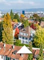 Multiple buildings in urban setting, brown roofs, green trees, cityscape view, mountains in background.