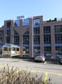 multi-story building, numerous glass windows, cars parked in front, rooftop greenery, gray exterior