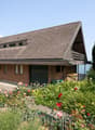 2-story brick house with a slanted roof, surrounded by a well-maintained garden with various flowers and plants. The house has a garage attached to the side.