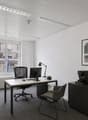 A modern office space with a desk, computer, and office chair. The room features a white wall, a radiator, and a window with a view of a building. There is a plant on the desk and a picture frame on the shelf. The room is empty and well-lit.