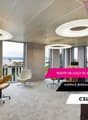 Large open office space with floor-to-ceiling windows offering a scenic view, modern furniture including round tables and swivel chairs, and colorful lighting fixtures on the ceiling.