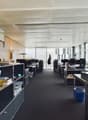Open office space with cubicles, desks, and storage units. The ceiling has exposed beams and the walls are mostly glass, allowing natural light to enter the space. There are various office equipment and supplies visible, including a dishwasher.