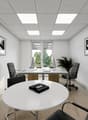 This image shows a modern and minimalist office space. The room has a white ceiling with recessed lighting panels, white walls, and a tiled floor. There is a large round white table in the center of the room, surrounded by black office chairs. Two framed