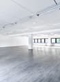 Large open office space with white walls, gray laminate flooring, and overhead fluorescent lighting. The space has large windows along one side providing natural light. The room appears to be empty and ready for use.