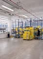 Large industrial warehouse space with shelving units, storage boxes, and a reception desk. The space has high ceilings, concrete floors, and overhead lighting.