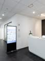 The image shows a modern office space with a reception area. The space has a clean, minimalist design with white walls, a gray floor, and a long white reception desk with a laptop on it. There is a large digital display screen in the center of the room, a