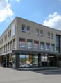 A 3 story building, grey walls, glass windows, signages, cars parked in front