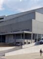 The building is made of concrete with a metallic look. It is on the corner of the street and has three floors with a basement. There is a ramp on the side.