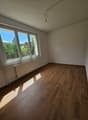 This image shows an empty room with hardwood flooring and white paneled walls. The room has large windows that provide a view of the surrounding greenery outside. The room appears to be a bedroom or living space with a simple and minimalist design.