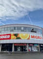 a modern, commercial building with storefronts, glass windows, and a curved facade. There are large advertisement boards with pictures of oranges on it.