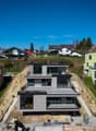 Modern house under construction, multiple levels, large windows, balconies, surrounded by other houses and greenery