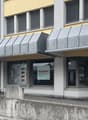 2 story building, yellow wall, grey roof, entrance with metal railings, glass windows, large awnings