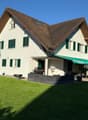 2-story house with a brown tiled roof, white exterior walls, and green shutters. The house has a covered patio or balcony area with a retractable awning. There is a garage and additional parking space on the property. The house is surrounded by a well-man
