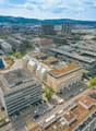 The image shows an aerial view of a dense urban cityscape with a mix of modern high-rise buildings and older structures. The city appears to be located in a hilly or mountainous area, with mountains visible in the background. The streets are lined with pa
