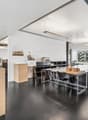Large open-plan office space with high ceilings, white walls, and concrete flooring. The space contains various workstations, desks, and chairs, as well as architectural models and displays on shelves. There is a view of the city through the large windows