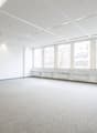 Large open office space with multiple windows providing a view, white walls, gray flooring, suspended ceiling with recessed lighting