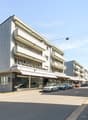 Multi-story apartment building with a modern, gray and white facade. The building has balconies and there are several parked cars visible in front of the building.