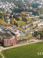 Large building with green roofs, solar panels, wide parking area, nearby roads, multiple smaller buildings, greenery, distant urban landscape