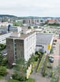 Aerial view of an urban area with buildings, parking lots, trees, and distant hills.