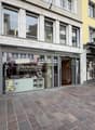 The storefront is a multi-story building with large windows, glass doors, and awnings. Inside the storefront, there is a shop with a display of products. The storefront is located in a busy street with other shops and a brick sidewalk.