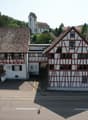 Building with a traditional design, two stories, brown and white checkered exterior, large windows, entrance with a staircase, sign reading 'Wurschaus Sormen', small wooden bench, church with a steeple in the background, green surroundings