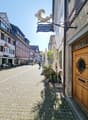 a cobblestone street, people walking, TAKE AWAY sign, outdoor dining tables, potted plants