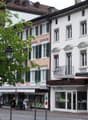 Three buildings with different colors, one with a balcony, shops on the ground floor, street lamp, tree, and bench