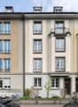 5-story building, beige and white exterior, multiple windows, balconies, front garden with plants and bicycles