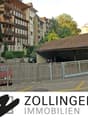 Building with a garage, a red car parked in front, a white car parked in the garage, a wooden fence, a building with balconies, and a tree in the background.