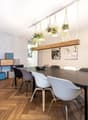 The image shows a modern and stylish dining room. The room has a large wooden dining table with black chairs around it. Above the table, there is a wooden beam with hanging plants in macrame planters, adding a touch of greenery to the space. The walls are