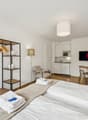 Compact bedroom with wooden floor, double bed, microwave, tv, dining table, small kitchen, hanging cabinet, floor lamp, and chair.