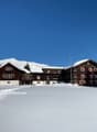 A wooden building covered in snow with several windows, two smaller houses on the left, trees in the front, mountains in the background